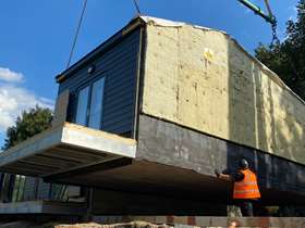 A modular house is lifted into place on a crane. There is a worker in an orange high vis vest helping with the task, which is taking place on a bright, sunny, blue sky day.